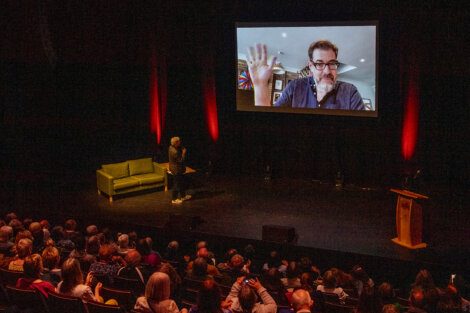 A person on stage gives a presentation to an audience, with a large screen displaying a video call of another person waving. The stage is dimly lit with red accent lights.