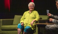 An elderly woman with short white hair, glasses, and a green shirt sits on a green sofa during an interview. She faces the interviewer, who holds papers and sits at a table with a glass of water.