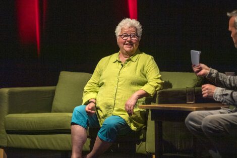 An elderly woman with short white hair, glasses, and a green shirt sits on a green sofa during an interview. She faces the interviewer, who holds papers and sits at a table with a glass of water.