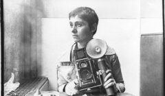 Black and white photo of a person with short hair sitting indoors, holding a vintage camera with a flash.