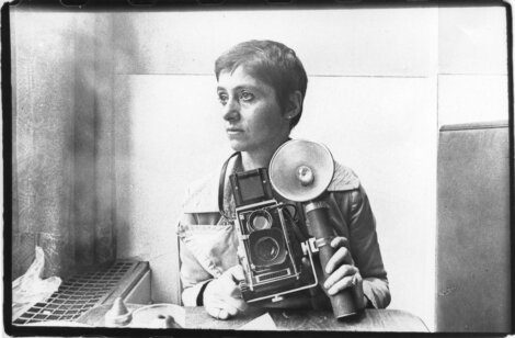 Black and white photo of a person with short hair sitting indoors, holding a vintage camera with a flash.