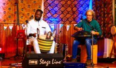 Two musicians perform on stage, one playing a traditional drum while the other plays a handpan. The backdrop features patterned tapestries, and various musical instruments are visible in the background.