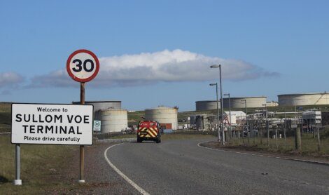 A road sign reads "Welcome to Sullom Voe Terminal. Please drive carefully" next to a 30 mph speed limit sign. Industrial tanks and a vehicle are visible in the background.