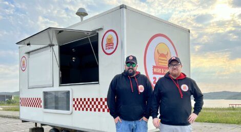 Two men wearing matching hoodies and caps stand in front of a white food truck with "Snack & Chat 2 Cows" logos on it. The food truck is parked outdoors with scenic landscape in the background.