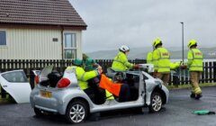 Firefighters in yellow jackets and helmets work at the scene of a car accident, with the roof of a silver car removed for rescue operations.