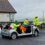 Firefighters in yellow jackets and helmets work at the scene of a car accident, with the roof of a silver car removed for rescue operations.