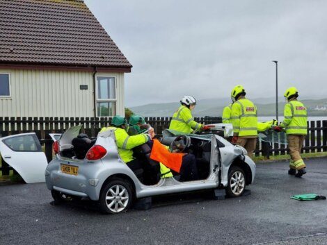 Firefighters in yellow jackets and helmets work at the scene of a car accident, with the roof of a silver car removed for rescue operations.