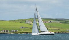 A sailboat with white sails navigates the water near a coastal area with green hills, buildings, and a few houses in the background under a partly cloudy sky.