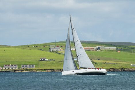 A sailboat with white sails navigates the water near a coastal area with green hills, buildings, and a few houses in the background under a partly cloudy sky.