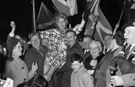 A jubilant crowd celebrates by lifting a smiling woman into the air. The scene is filled with waving flags and happy faces.