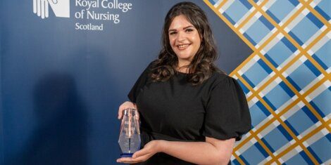 A person in a black dress holding an award stands in front of a backdrop that reads "Royal College of Nursing Scotland.