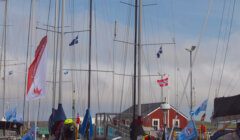 Sailboats with various flags docked at a marina. A red building with a peaked roof is in the background against a partly cloudy sky.