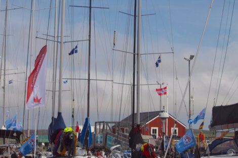 Sailboats with various flags docked at a marina. A red building with a peaked roof is in the background against a partly cloudy sky.