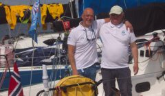 Two men in white shirts stand arm-in-arm on a docked sailboat with Norwegian flags and yellow jackets visible in the background.