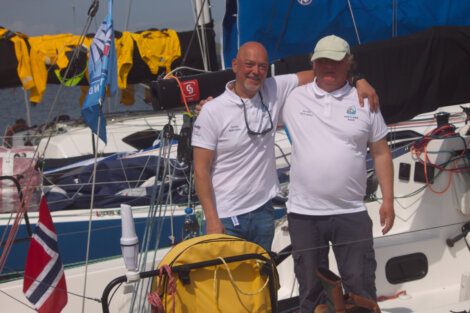 Two men in white shirts stand arm-in-arm on a docked sailboat with Norwegian flags and yellow jackets visible in the background.