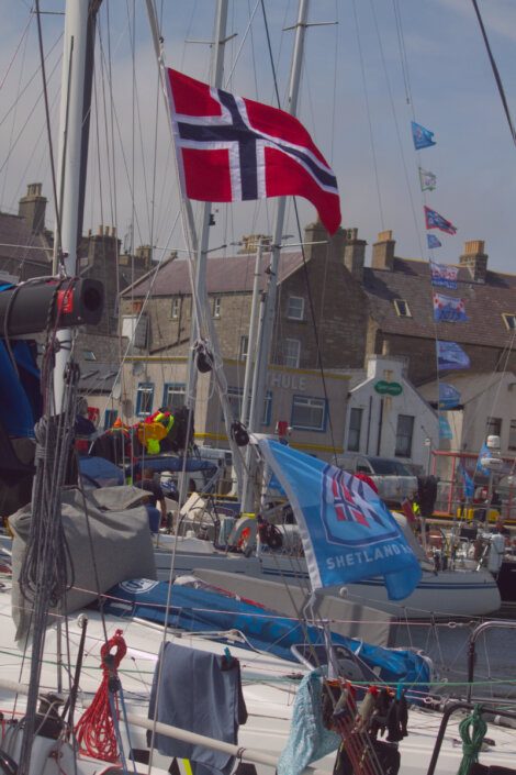 A docked boat with Norwegian and Shetland flags. Buildings and people are visible in the background.