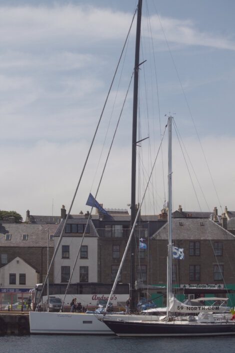 A sailboat docked in a harbor with buildings and shops in the background under a partly cloudy sky.