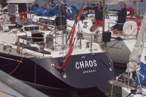 A sailboat named "Chaos" from Arendal is docked at a marina. Several people are on and around the boat, and other boats are nearby.