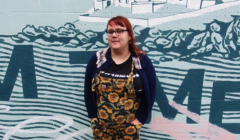 A person stands in front of a mural. They are wearing eyeglasses, a blue cardigan, and sunflower-patterned overalls with hands in their pockets. The mural features abstract designs and a building outline.