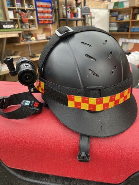 A black safety helmet with a red and yellow checkered band, equipped with a mounted camera, sits on a red surface in a workshop filled with various tools and equipment.