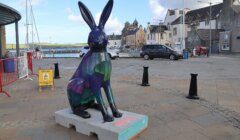 A colorful rabbit statue sits on a pedestal in a paved town square with buildings and cars in the background.