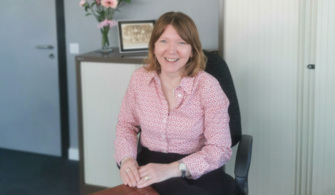 A woman with shoulder-length light brown hair sits at a desk, smiling. A vase of flowers and a framed photo are on the cabinet behind her.