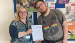 Two people standing in front of a bulletin board. The person on the left holds a certificate with the person on the right. The person on the right wears a t-shirt with a bear print.