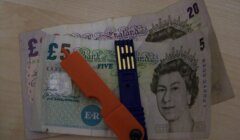 Three British banknotes - £5, £10, and £20 - placed on a table, with an orange and blue USB drive on top of them.