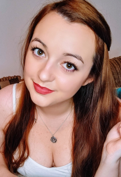 A woman with long brown hair and light makeup smiles at the camera, wearing a white sleeveless top and a necklace with a circular pendant. Brownish-gray background.