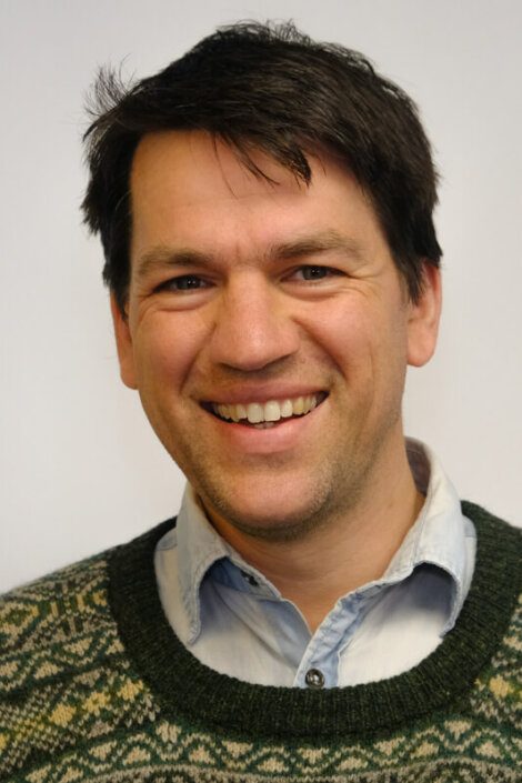 A man with short dark hair smiles at the camera. He is wearing a light blue collared shirt under a green and beige patterned sweater. The background is plain white.