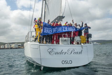 A group of people on a sailing yacht named "Ender Pearl" holding a blue banner that reads "Bergen Shetland Blue Ribbon." Buoyant celebration with fists raised. The boat is docked with an industrial background.