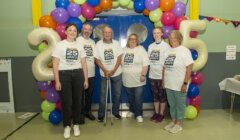 Six people wearing matching "25 years" t-shirts stand in front of a colorful balloon arch with the number 25.