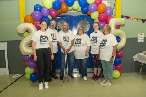 Six people wearing matching "25 years" t-shirts stand in front of a colorful balloon arch with the number 25.