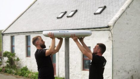 Two men are installing a Kymeta satellite dish on a pole in front of a white building with a pitched roof.