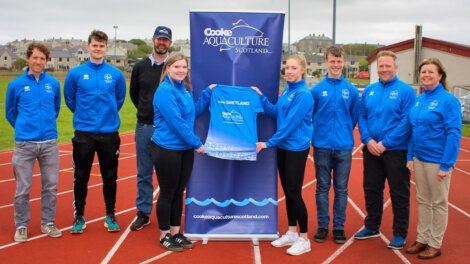 A group of people in blue jackets stand on a track, presenting a T-shirt in front of a "Cooke Aquaculture Scotland" banner.