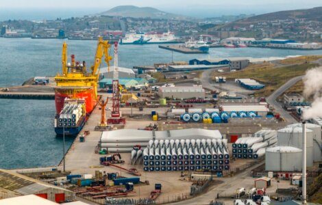 A port with a large ship docked, surrounded by wind turbine components, cranes, storage tanks, and industrial buildings. Hills and a distant town are visible in the background.