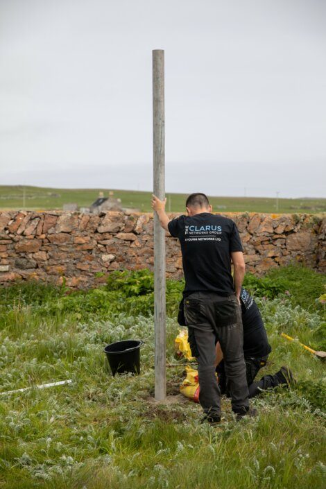 Two individuals wearing black shirts are installing a pole in a grassy field. One person is holding the pole, and the other is bending down, working with tools. A stone wall is visible in the background.