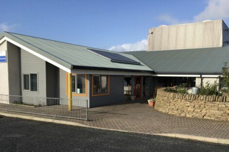 A modern, single-story building with a metal roof and solar panel, features large windows, an entrance with red-framed doors, and a stone wall bordering a paved walkway.