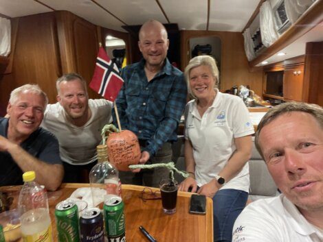 Five people smiling and posing around a table on a boat, with beverages and a pumpkin in the center. One person is holding a Norwegian flag.