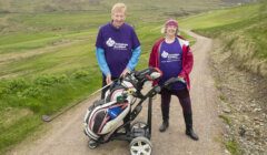 Two people wearing purple Alzheimer Scotland t-shirts stand on a golf course with a golf trolley and bags, surrounded by grassy hills and pathways.