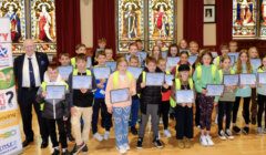 A group of children and an adult pose with certificates in front of a banner about road safety. Stained glass windows are in the background.