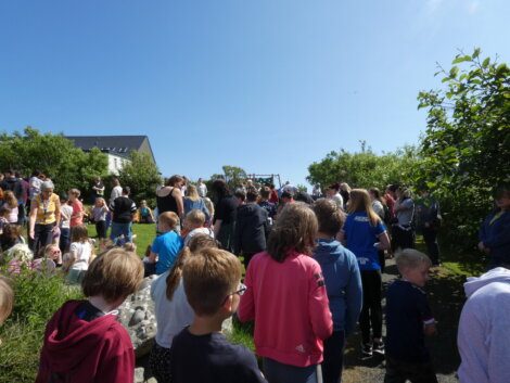 A crowd of people, including children and adults, standing outdoors on a sunny day with blue skies. Trees and a building are visible in the background.