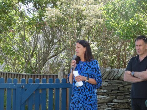 A woman in a blue and black patterned dress speaks into a microphone outdoors next to a man in a black shirt, with trees and a stone fence in the background.