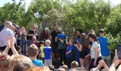 A group of people, including children and adults, gather outside in a garden. One person in blue holds a covered item, while others clap and watch. Trees and bushes surround the group.