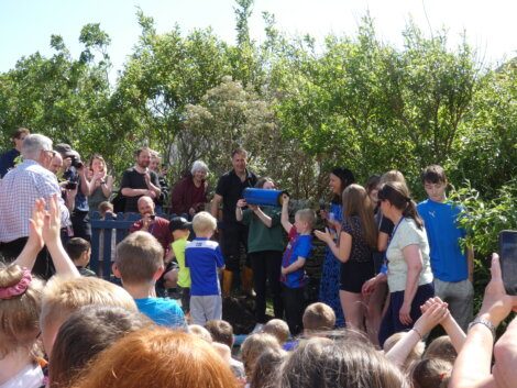 A group of people, including children and adults, gather outside in a garden. One person in blue holds a covered item, while others clap and watch. Trees and bushes surround the group.