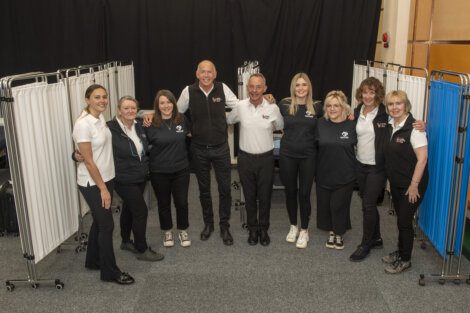 A group of nine people, some wearing matching uniforms, stand inside a medical screening area, smiling for a group photo. They are positioned in front of privacy screens.