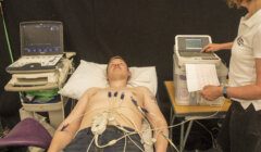 A man lies on a medical bed connected to various electrodes while a healthcare professional analyzes data from a monitor.