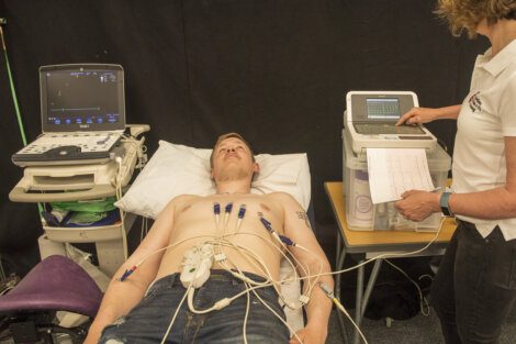 A man lies on a medical bed connected to various electrodes while a healthcare professional analyzes data from a monitor.