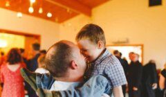 An adult man is holding a smiling boy in a room filled with people. The two are making eye contact, and the boy is wearing a checkered shirt and jeans.