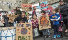 A group of people, including adults and children, hold signs advocating for environmental conservation. Signs include messages like "Save Our Planet," featuring a globe and flames.